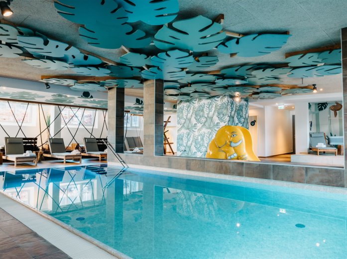 Innenpool mit Liegestühlen, blauen Blattformen an der Decke und gelbem Elefantenrutsch