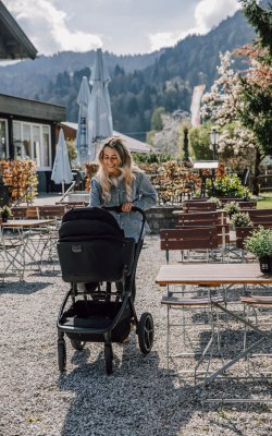 Frau schiebt Kinderwagen auf Kiesplatz neben Tischen und Stühlen im Freien mit Bergen im Hintergrund