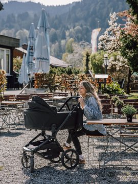 Frau sitzt an einem Tisch im Freien neben einem Kinderwagen in einem Garten mit Bergen im Hintergrund