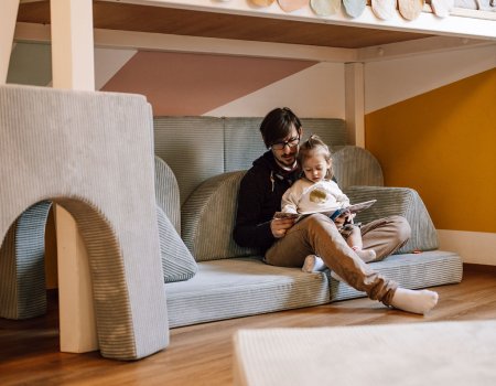 Vater liest mit Kind auf grauem Sofa unter einem Hochbett mit bunter Wimpelkette