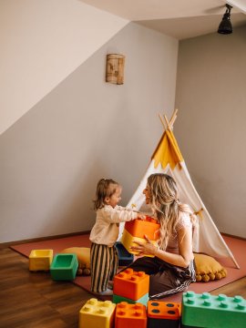 Kind und Frau spielen mit großen bunten Bausteinen vor einem weißen Tipi im Zimmer