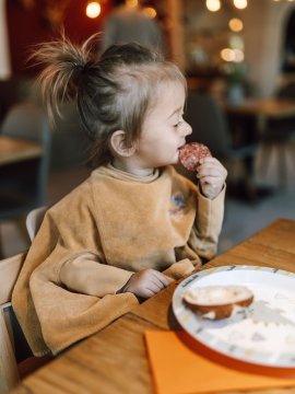 Child sitting at table eating a slice of sausage, plate with bread piece in front